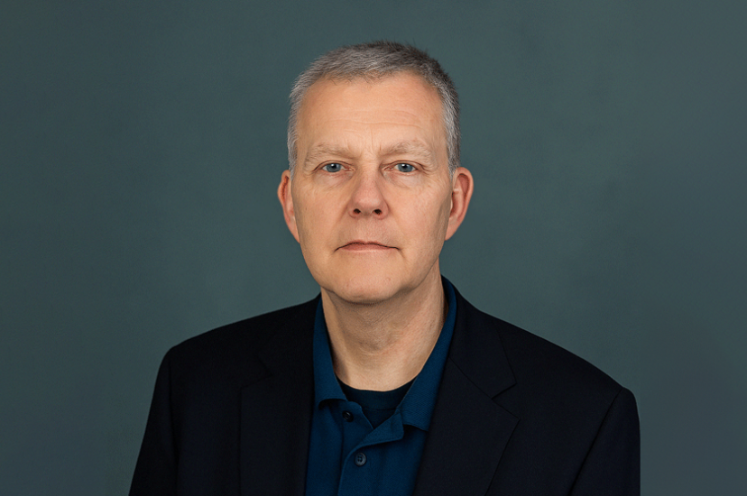 A man with salt and pepper hair, wearing a blue shirt and a dark jacket, sits in front of a green background