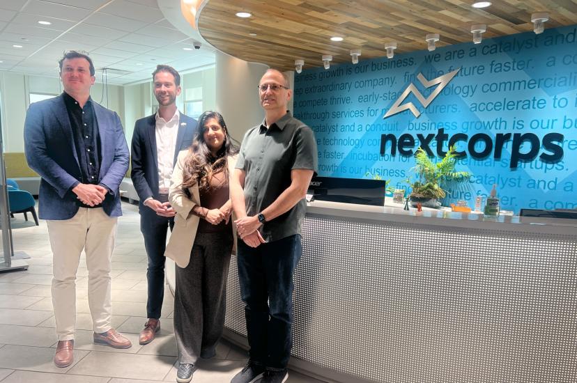 Three menand a woman standin front of a reception desk. The sign behind the desk says Nextcorps