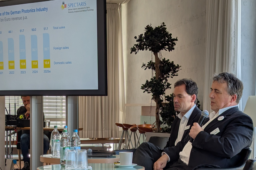 Two men in dark suits sit in front of a presentation on German photonics