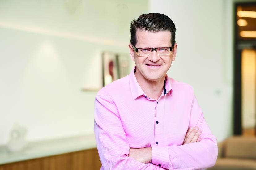 A man with brown hair and glasses, wearing a pink shirt, stands in front of a white wall 