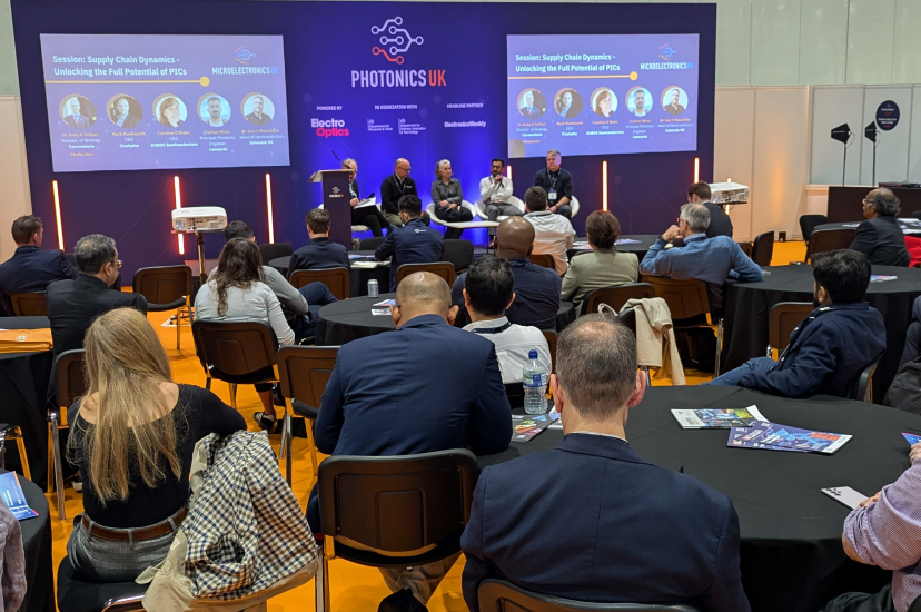 A crowd of people listening to four speakers in a panel discussion on a blue stage. The text on the backdrop reads PhotonicsUK