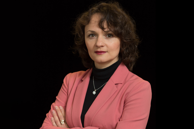 A white woman with curly auburn hair and wearing a salmon-coloured jacket, stands in front of a black background