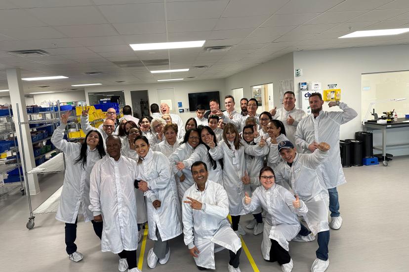 A large group of people wearing white lab coats and shoe covers are posing and smiling in a production or clean room environment