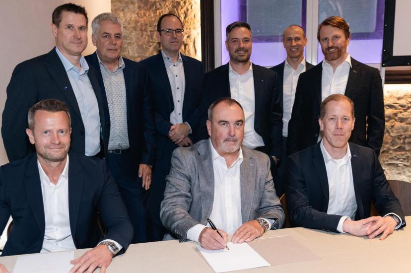 Three suited men sign contracts while sitting down at a table, with six more suited men stood behind