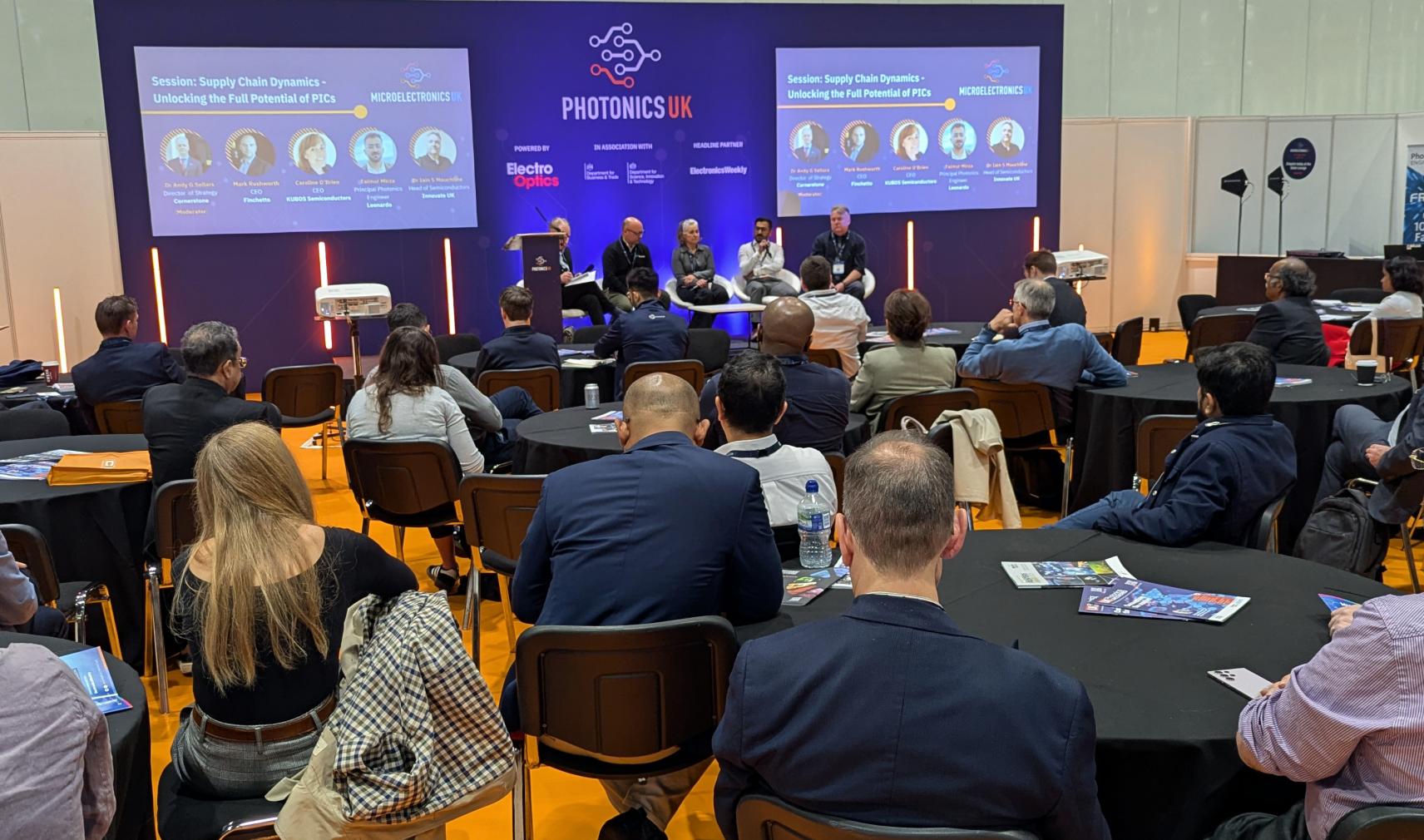 A crowd of people listening to four speakers in a panel discussion on a blue stage. The text on the backdrop reads PhotonicsUK