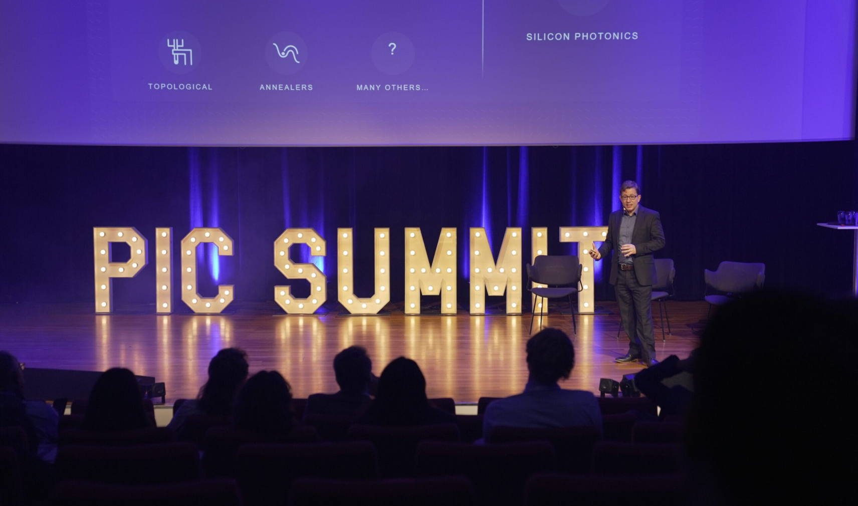 A man stands on a stage before a crowd. The words 'PIC SUMMIT' appear behind him on a purple backdrop