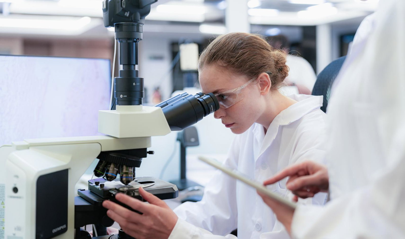 Team of female scientists collaborating in digital pathology lab. Young researcher adjusting microscope while colleague observes with tablet for medical diagnosis