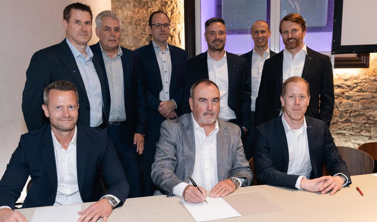 Three suited men sign contracts while sitting down at a table, with six more suited men stood behind
