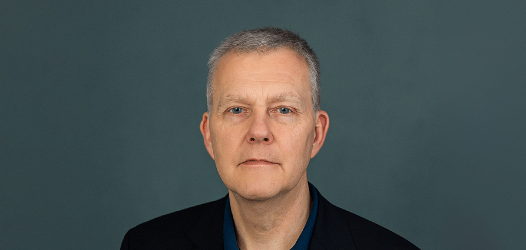 A man with salt and pepper hair, wearing a blue shirt and a dark jacket, sits in front of a green background