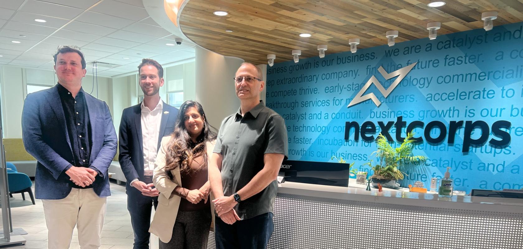 Three menand a woman standin front of a reception desk. The sign behind the desk says Nextcorps