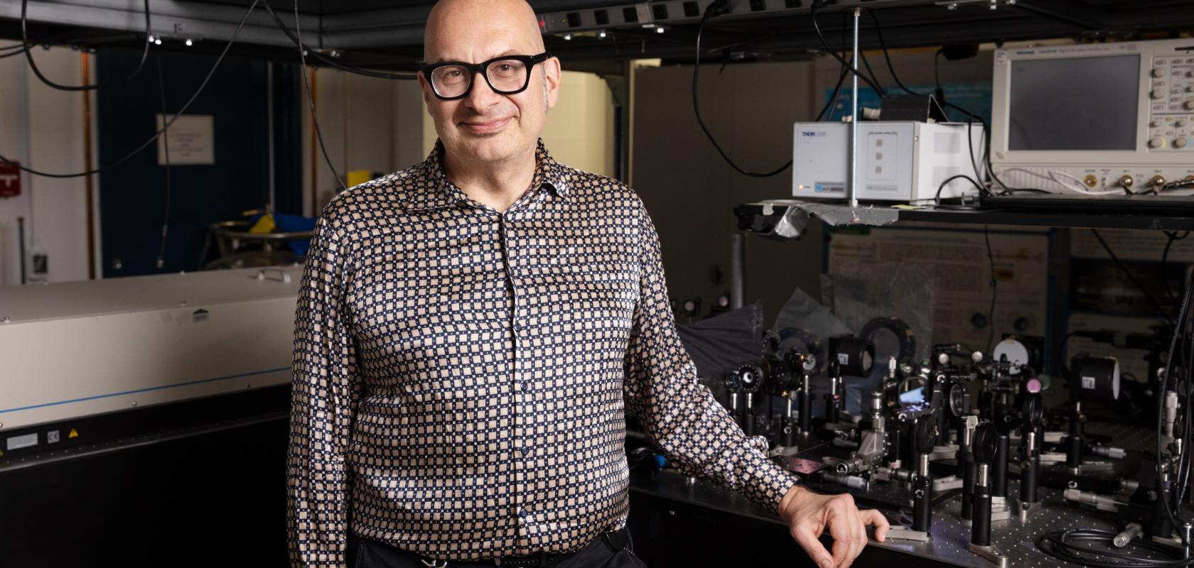 A bald man with glasses stands at an electronics bench