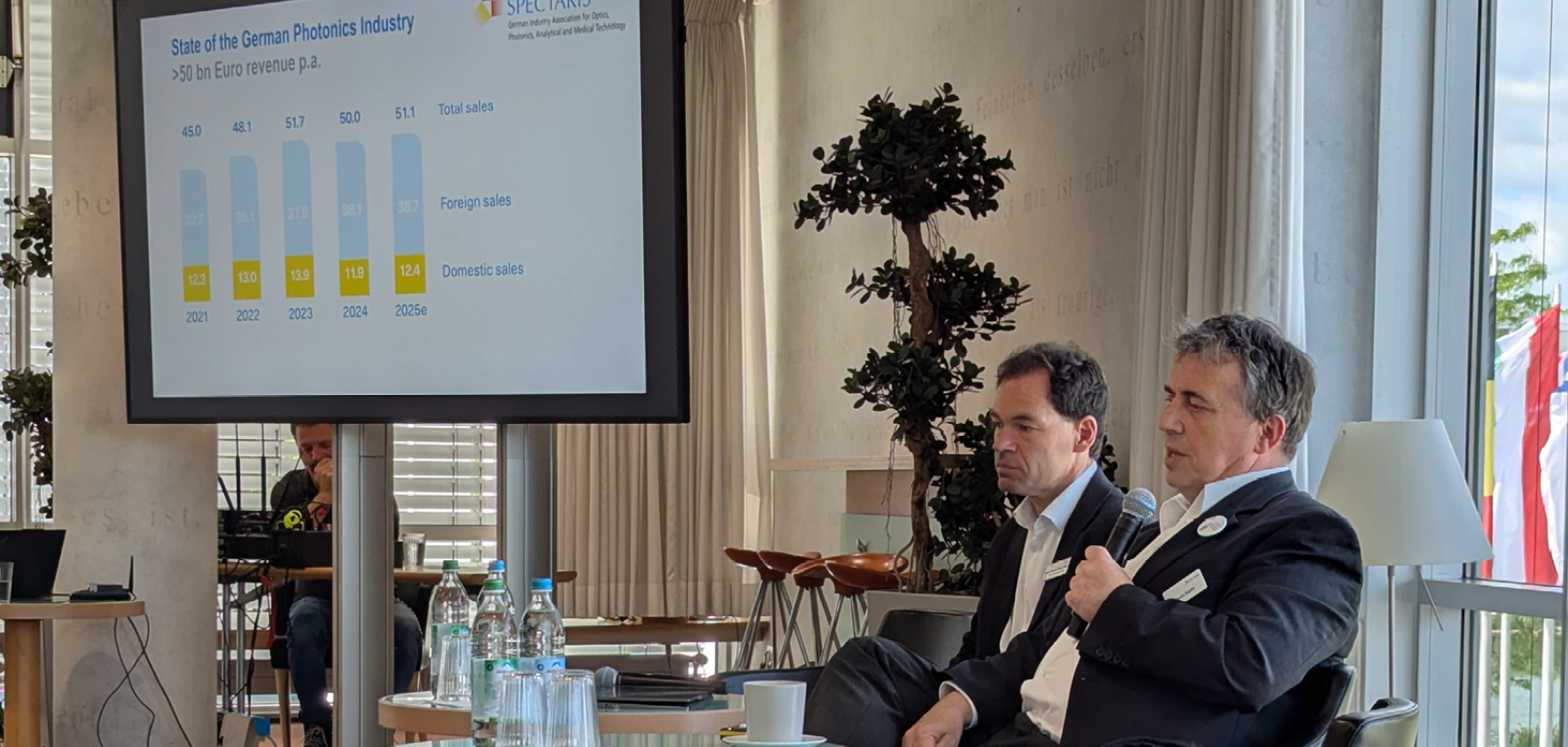 Two men in dark suits sit in front of a presentation on German photonics