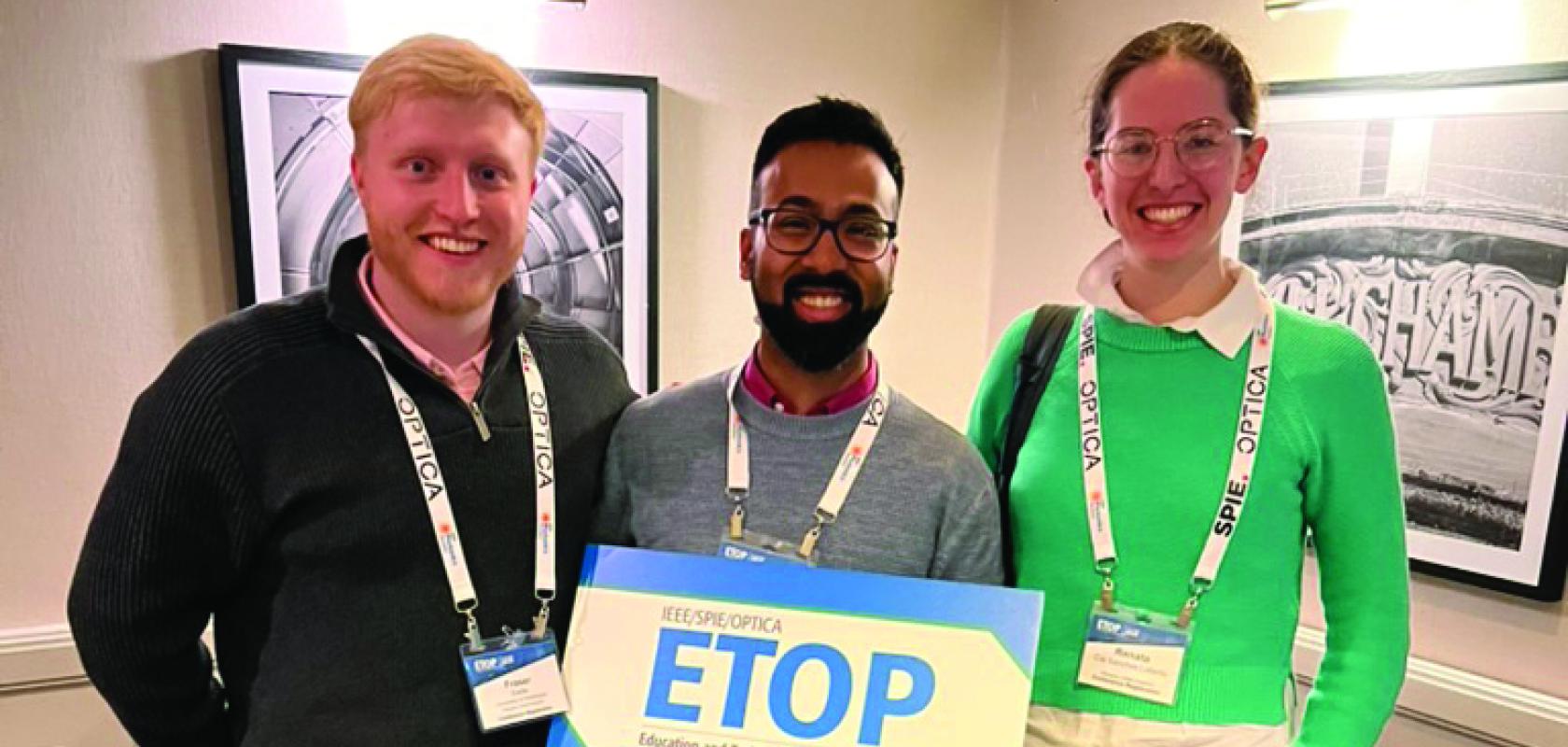 A fair-haired man, a dark-haired man with a beard and glasses, and a woman with light brown hair and glasses, hold a sign in a hotel corridor that says ETOP in blue letters