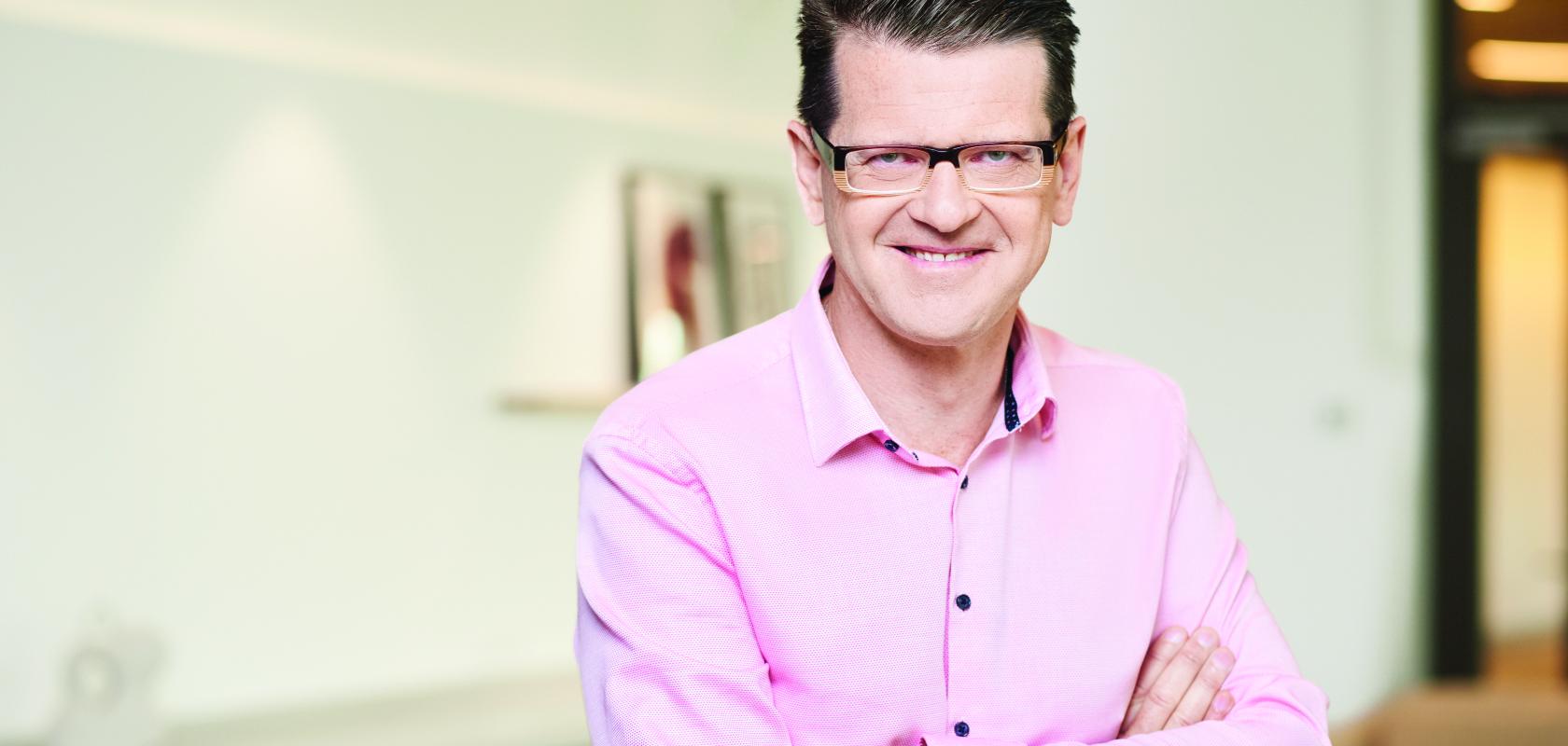 A man with brown hair and glasses, wearing a pink shirt, stands in front of a white wall 