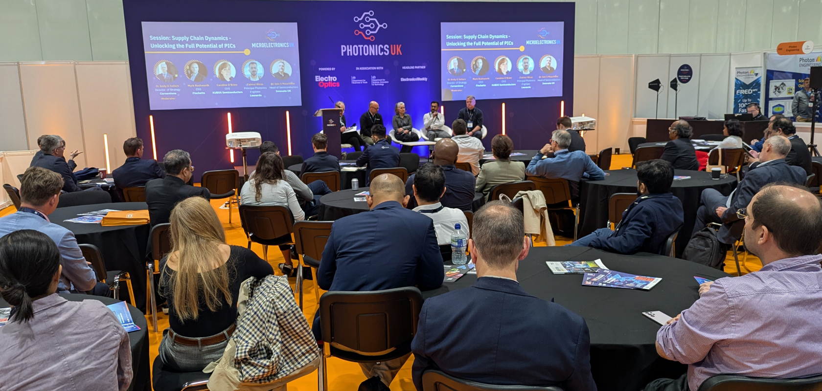 A crowd of people listening to four speakers in a panel discussion on a blue stage. The text on the backdrop reads PhotonicsUK