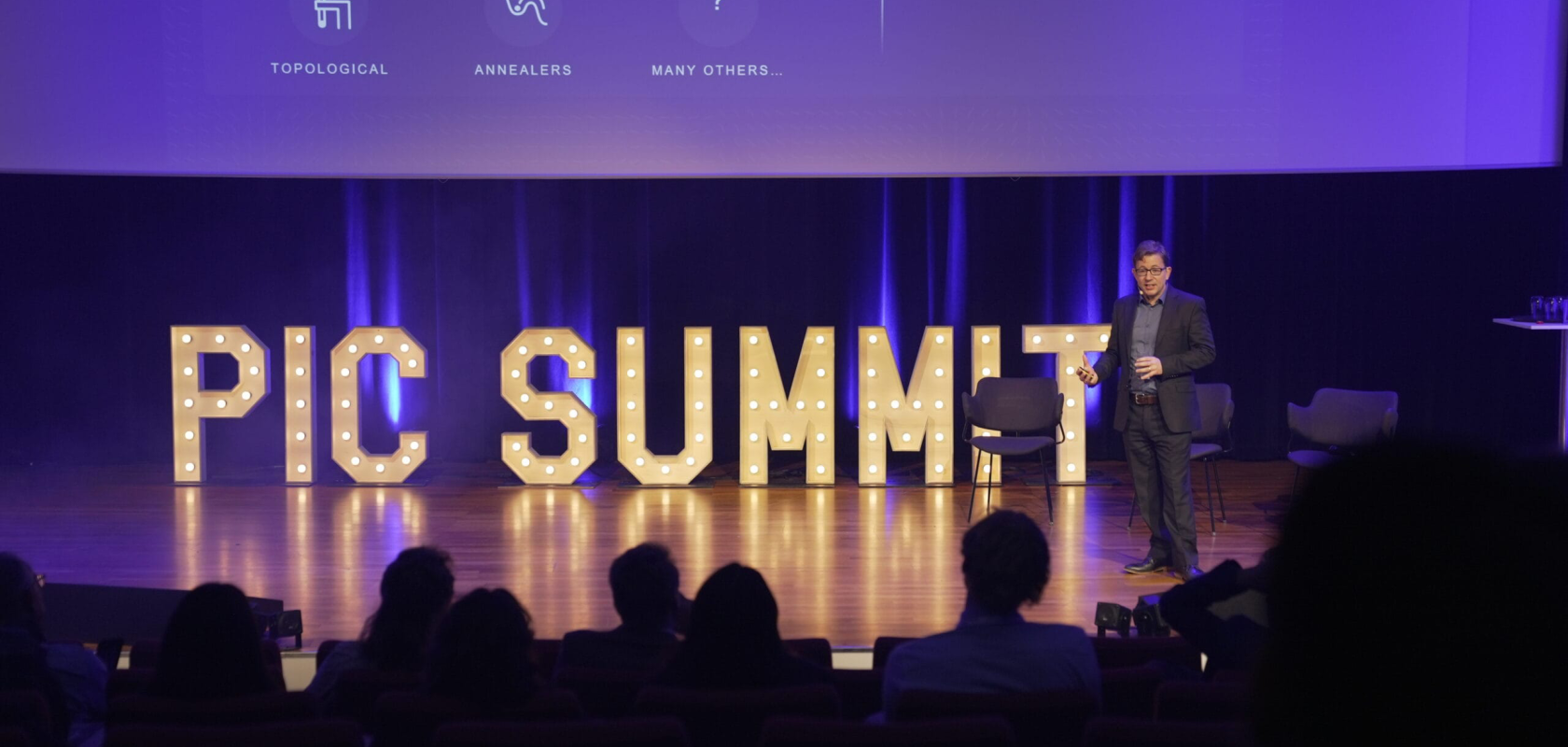 A man stands on a stage before a crowd. The words 'PIC SUMMIT' appear behind him on a purple backdrop