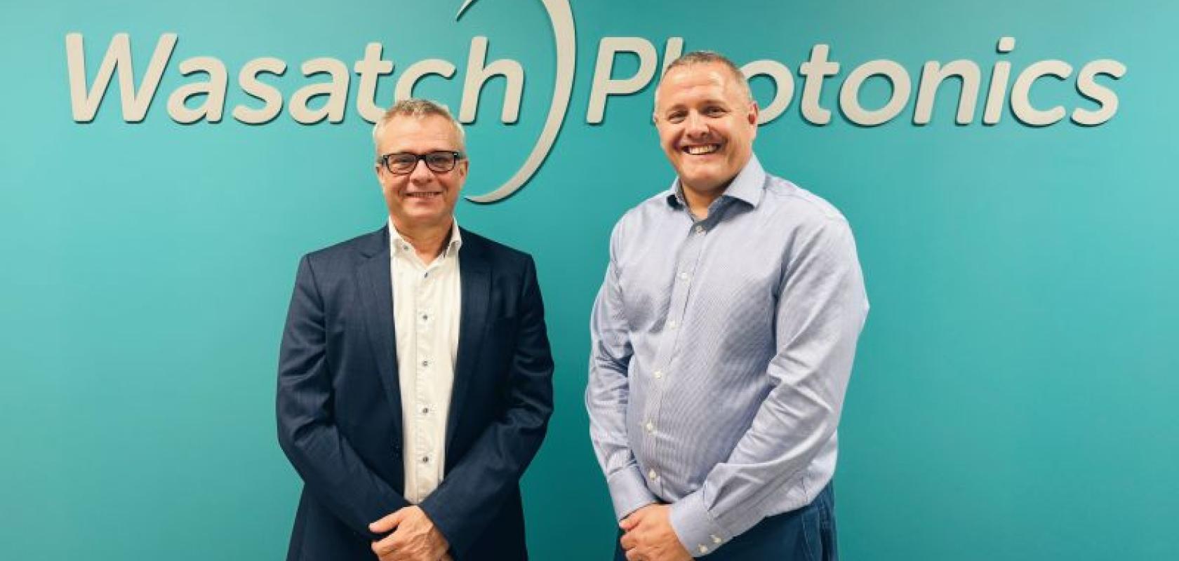 Two white men stand in front of a green wall. The words on the wll read Wasatch Photonics