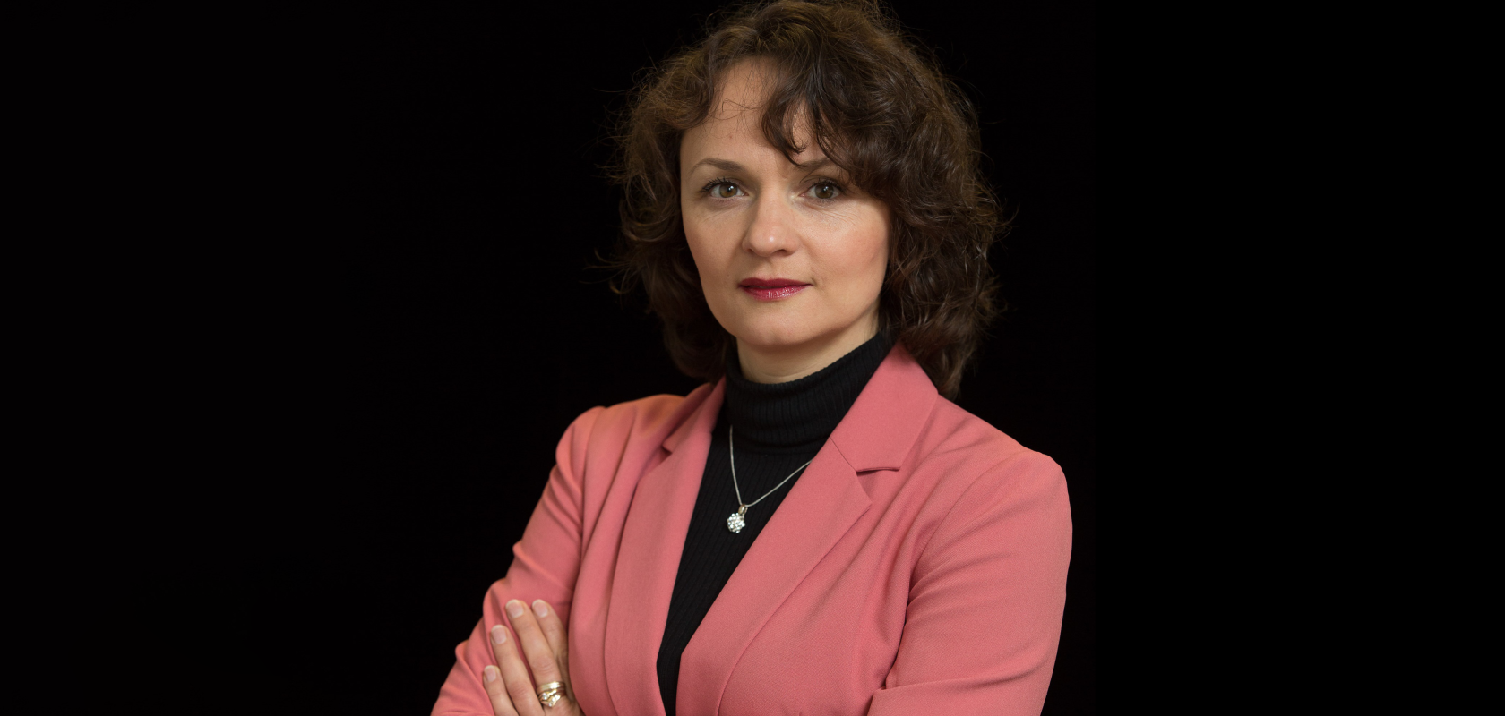 A white woman with curly auburn hair and wearing a salmon-coloured jacket, stands in front of a black background