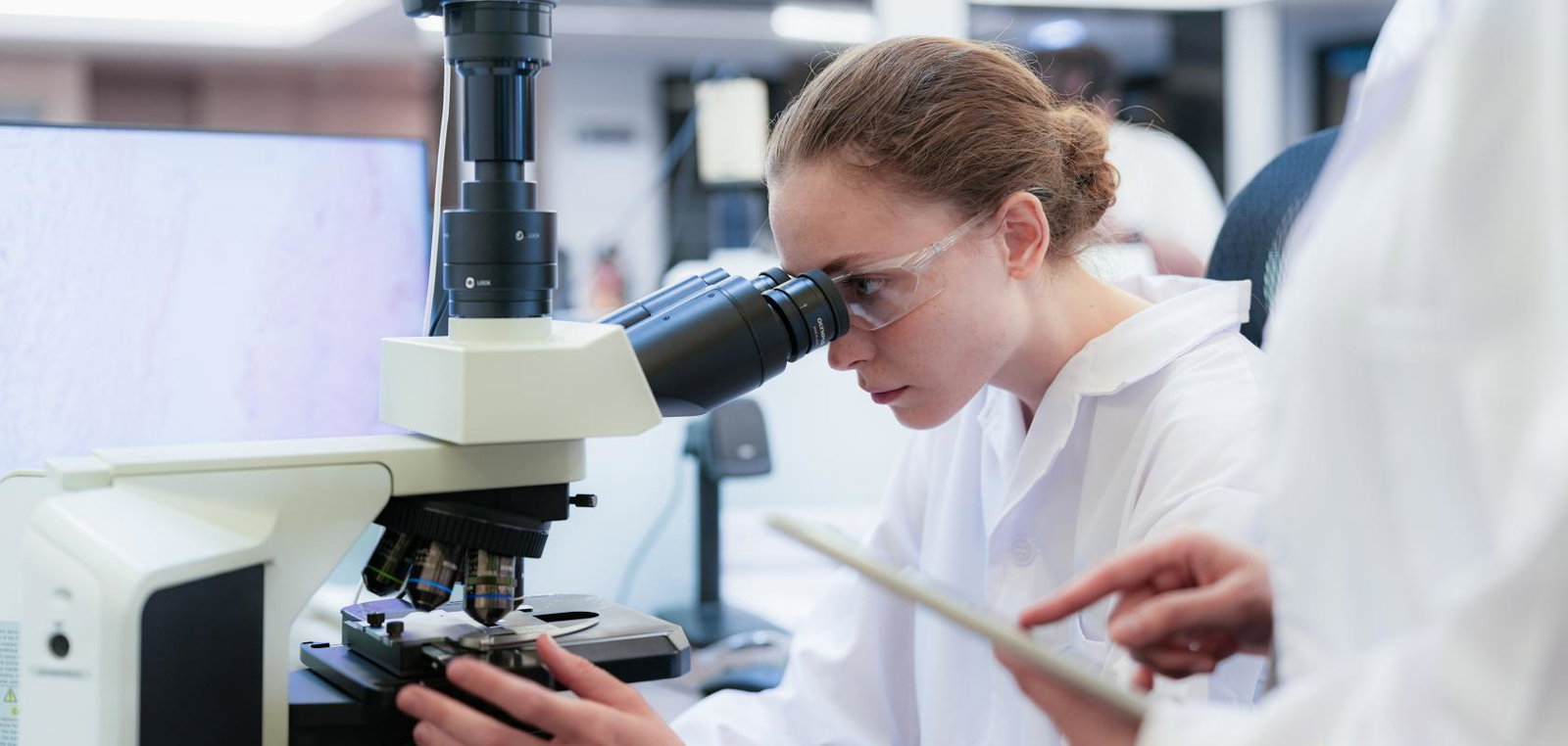 Team of female scientists collaborating in digital pathology lab. Young researcher adjusting microscope while colleague observes with tablet for medical diagnosis