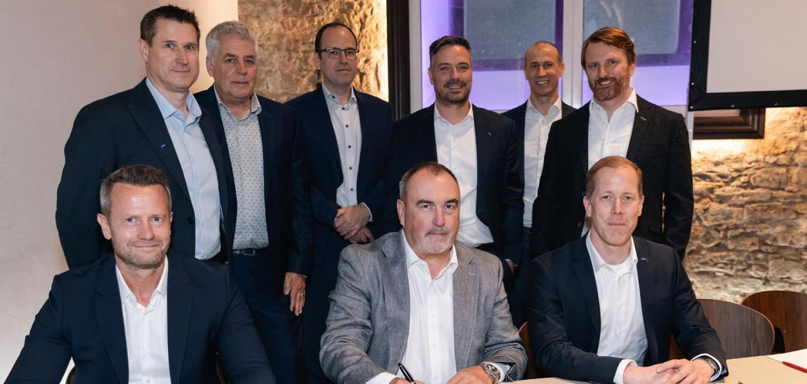 Three suited men sign contracts while sitting down at a table, with six more suited men stood behind