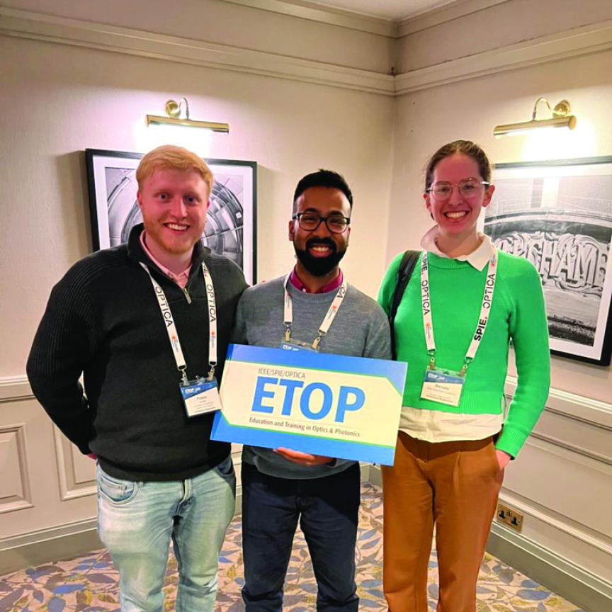 A fair-haired man, a dark-haired man with a beard and glasses, and a woman with light brown hair and glasses, hold a sign in a hotel corridor that says ETOP in blue letters