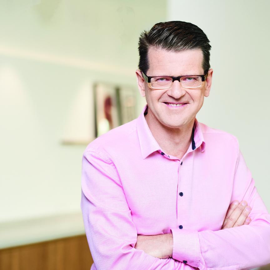 A man with brown hair and glasses, wearing a pink shirt, stands in front of a white wall 