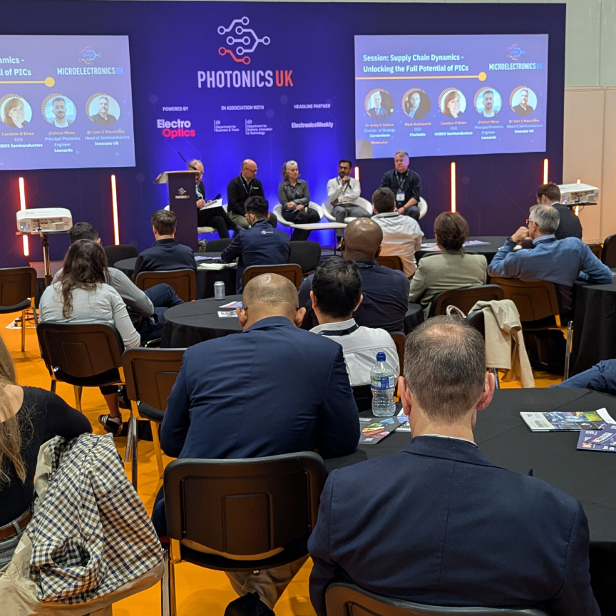 A crowd of people listening to four speakers in a panel discussion on a blue stage. The text on the backdrop reads PhotonicsUK