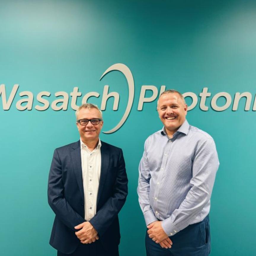 Two white men stand in front of a green wall. The words on the wll read Wasatch Photonics