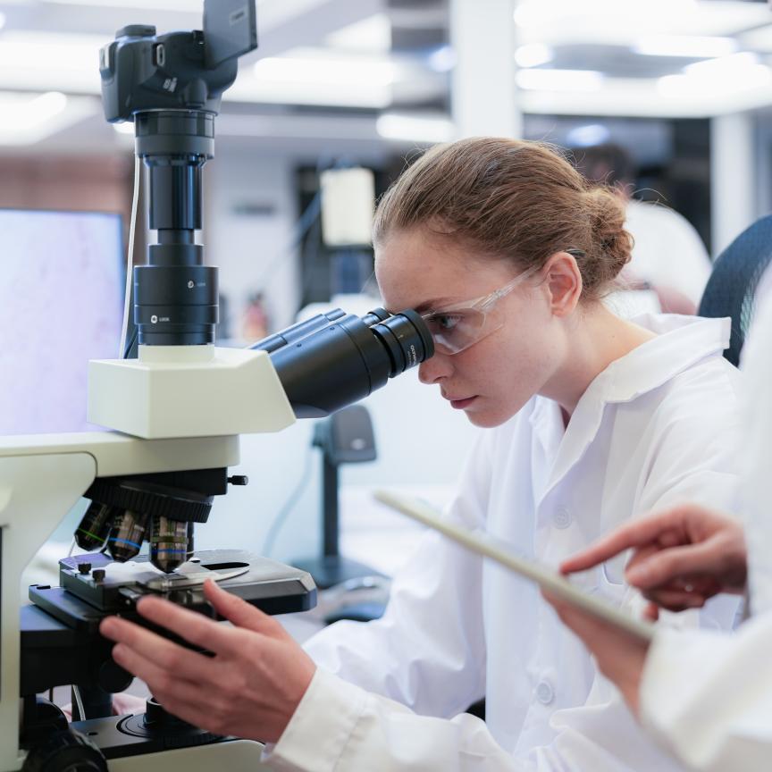 Team of female scientists collaborating in digital pathology lab. Young researcher adjusting microscope while colleague observes with tablet for medical diagnosis