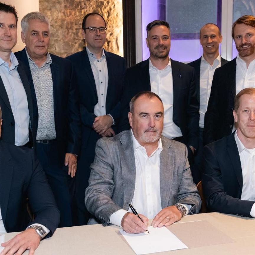 Three suited men sign contracts while sitting down at a table, with six more suited men stood behind